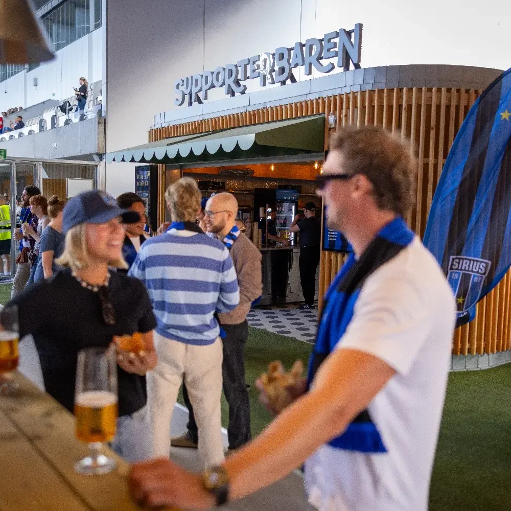 Personer som dricker öl framför Supporterbaren på Studneternas. Läktaren i bakgrunden.